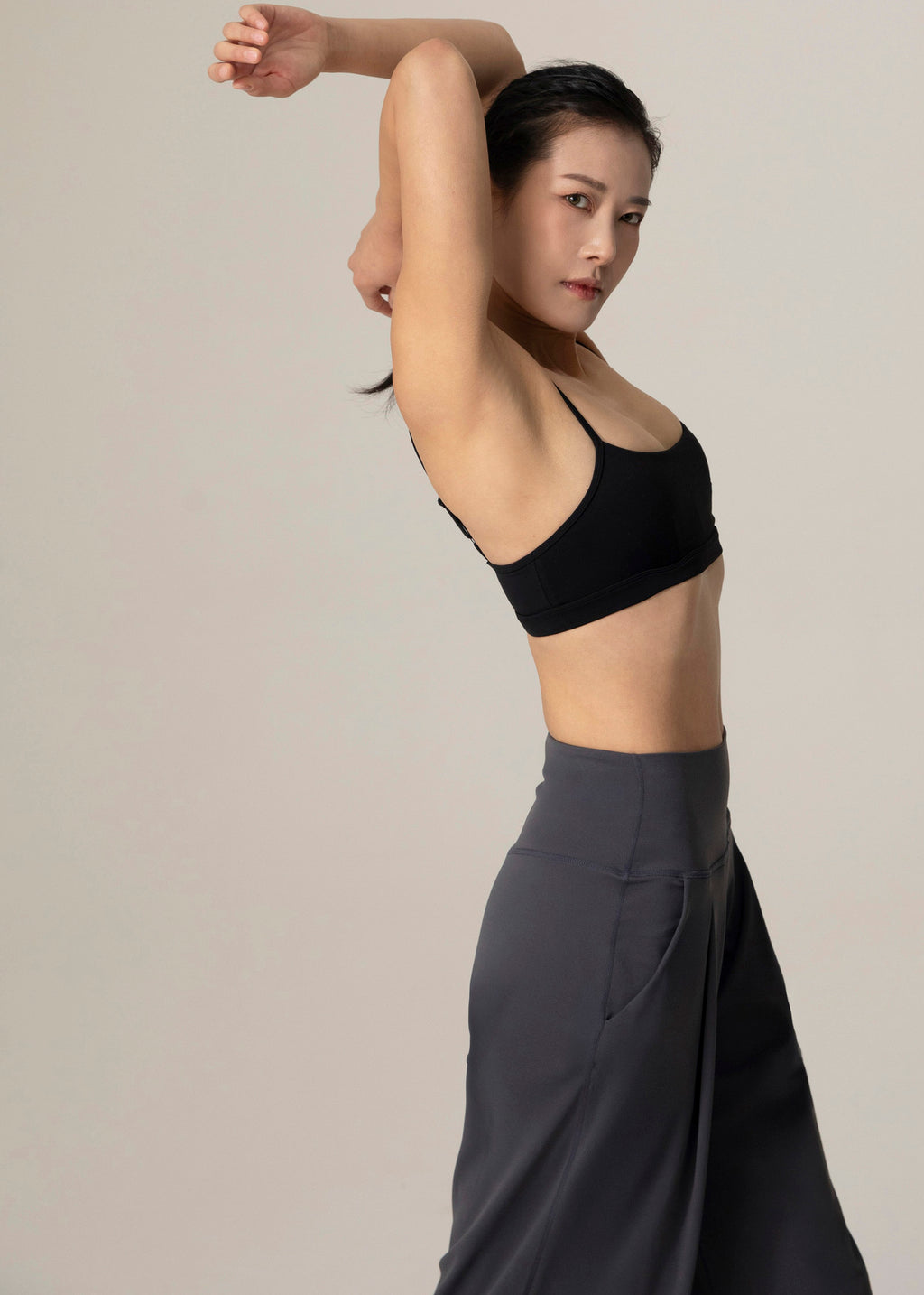 Closer side view of model wearing slate gray Flowmango Deep Flow wide-leg pants and black sports bra with arms bent overhead
