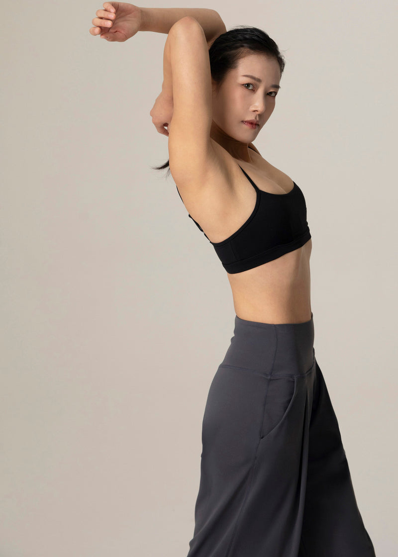 Closer side view of model wearing slate gray Flowmango Deep Flow wide-leg pants and black sports bra with arms bent overhead