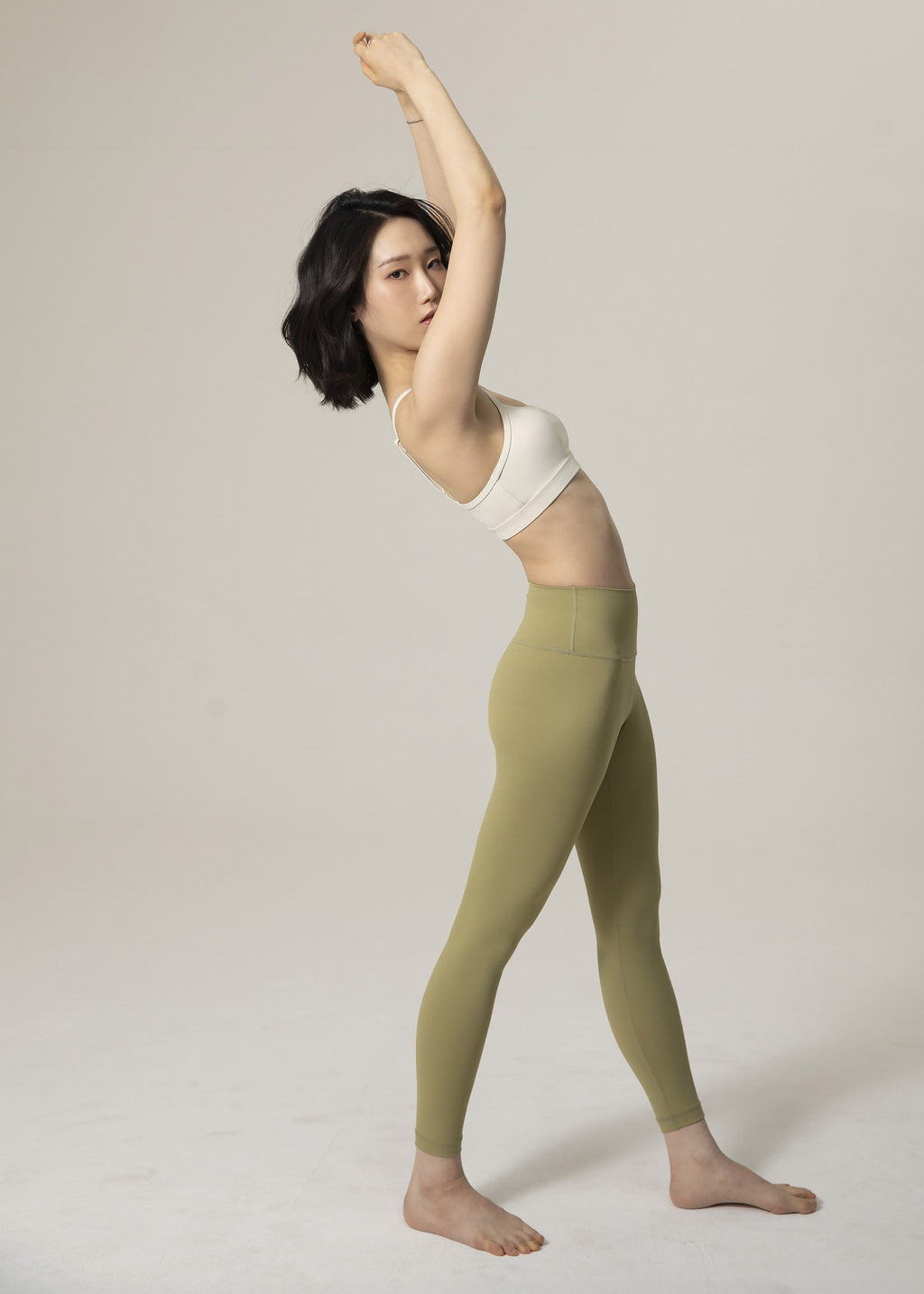 Side profile of model standing in olive green Flowmango Inner Flow leggings and ivory bra with arms raised
