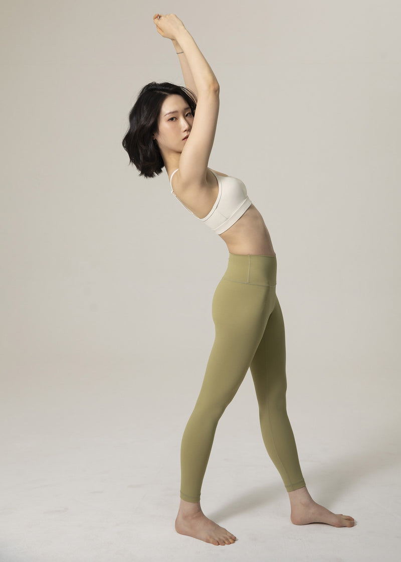 Side profile of model standing in olive green Flowmango Inner Flow leggings and ivory bra with arms raised