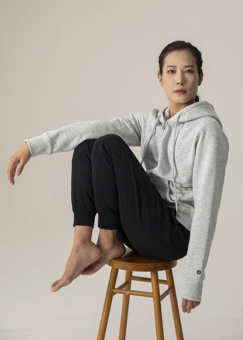 Model seated on a wooden stool in the heather gray Dot Flow hoodie and black jogger pants with one knee raised