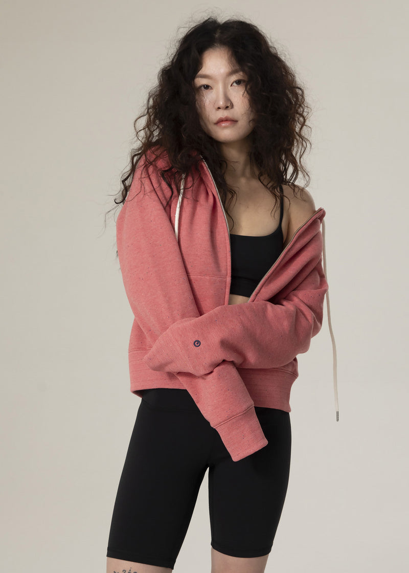 Model wearing the dusty rose Dot Flow zip-up hoodie off one shoulder over a black sports bra and bike shorts
